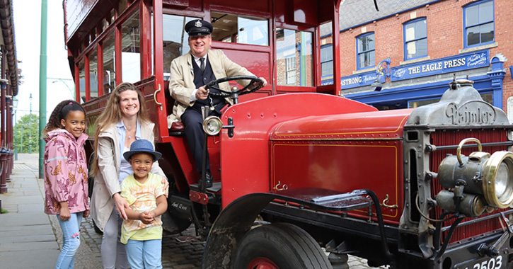 Family of three (woman and two children) smiling at camera next to a vintage red bus with bus driver sat behind driver's wheel.